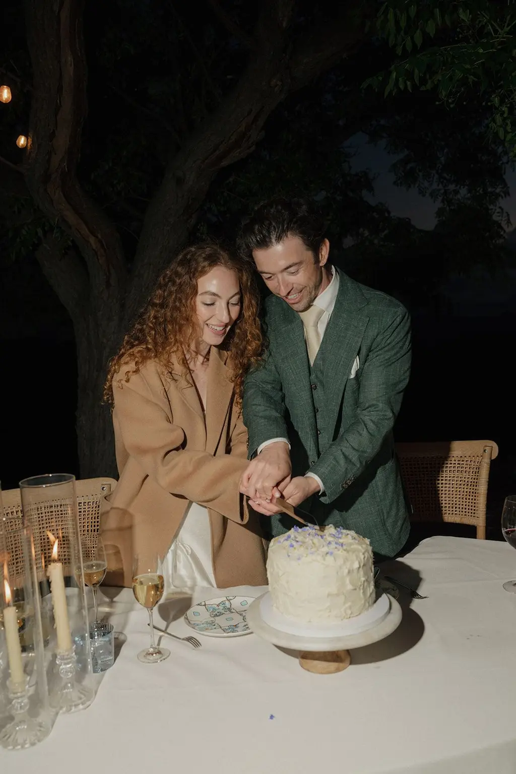 The couple cutting a small white cake by candlelight beneath the trees.