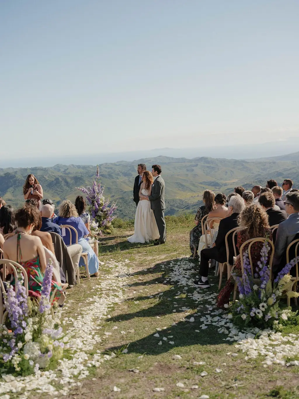 The couple at the end of a petal-strewn aisle, guests seated on either side, the ocean on the horizon behind them.