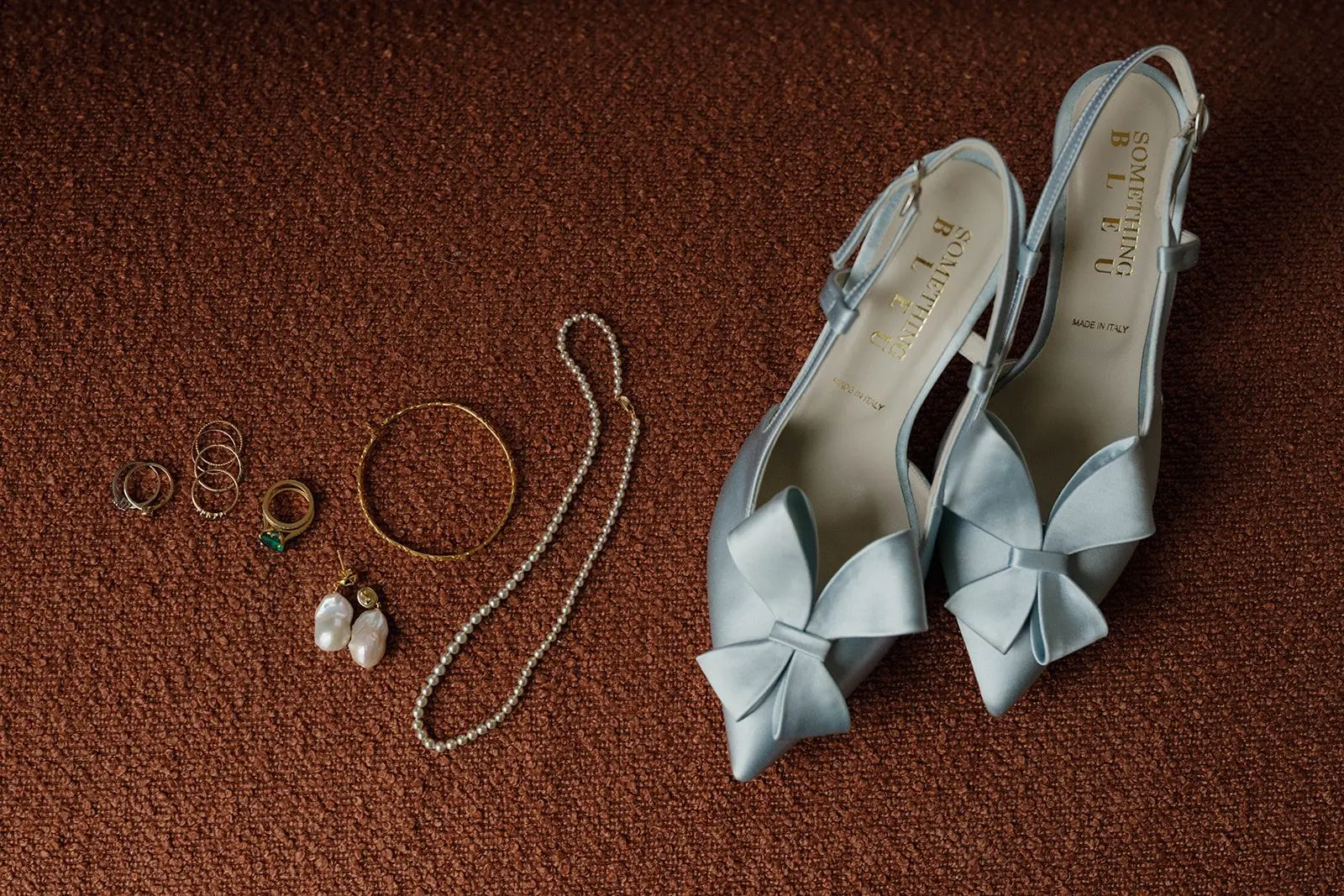 Pale blue satin bow-front heels laid out on rust-colored bouclé beside gold rings, pearl earrings and a pearl necklace.