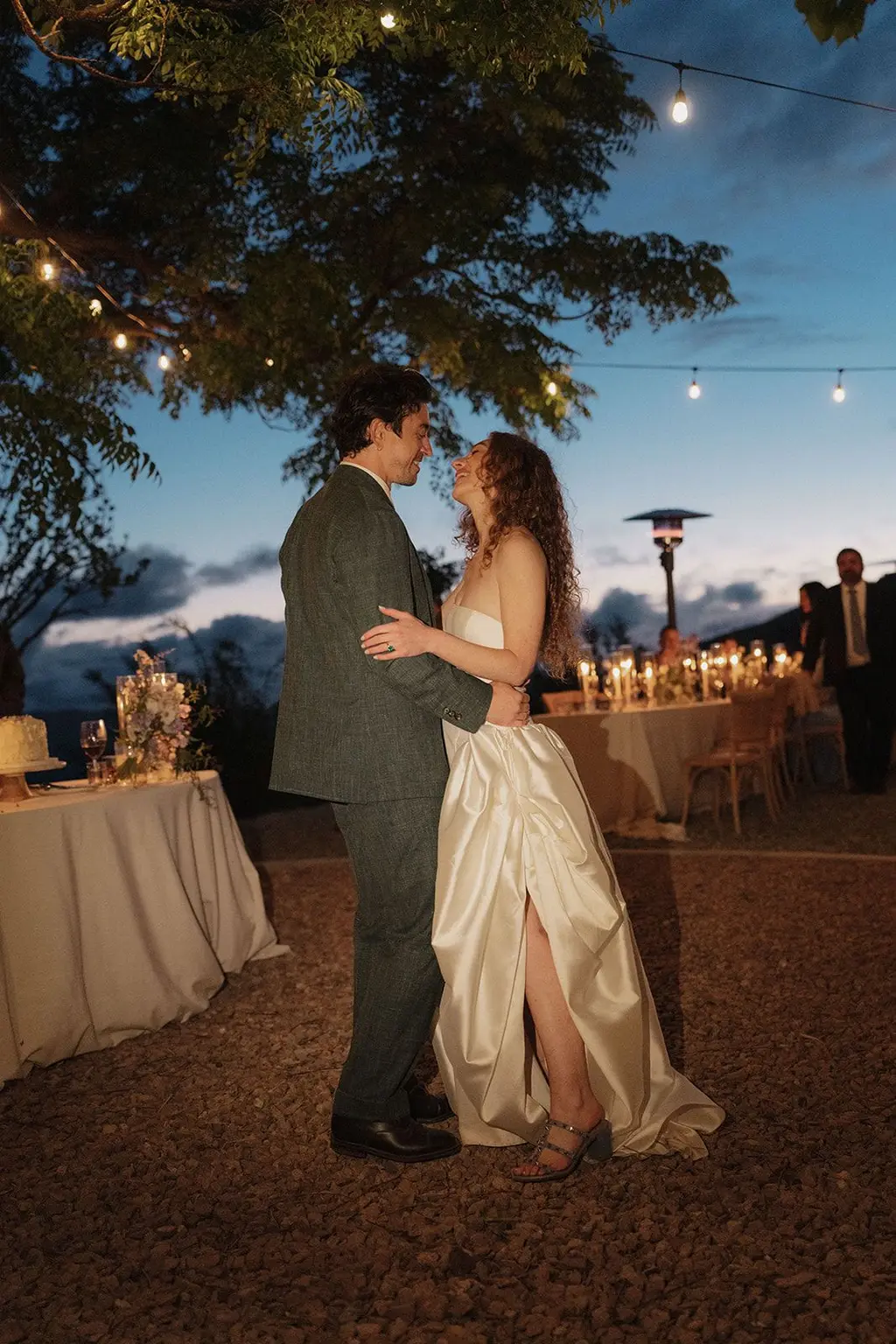 The couple dancing under strings of warm bulbs at blue hour, candlelit tables and the dark ridge behind them.