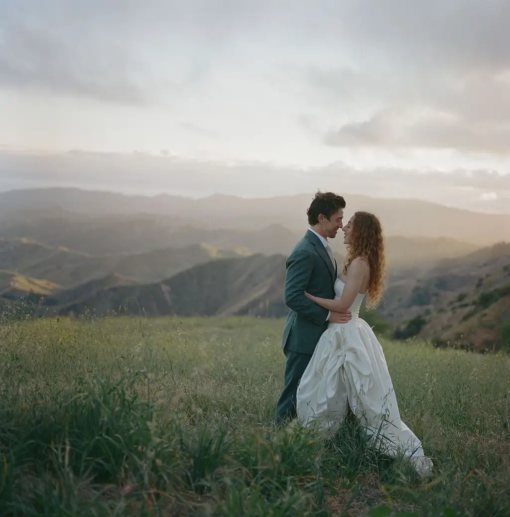 The couple in long grass at sunset, shot on film, the hills behind them washed in low gold light.