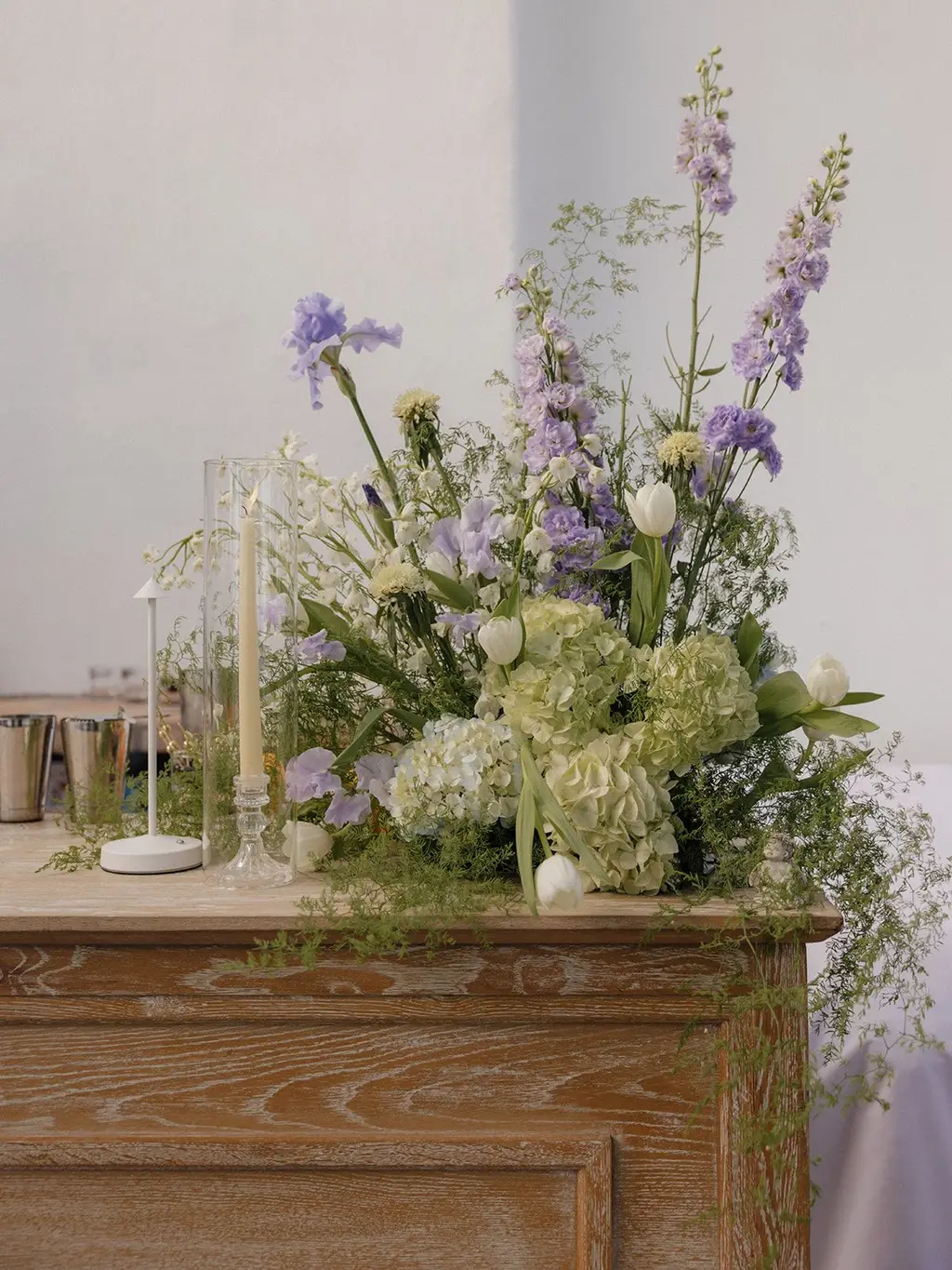 A loose arrangement of lavender iris, delphinium and white ranunculus on an oak sideboard beside lit tapers.